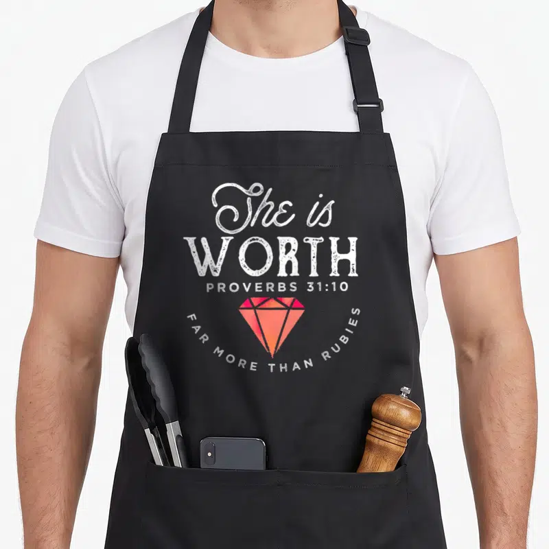 She the ruby apron - White stacked text and red orange diamond graphic on black bib apron — shown on a black cotton canvas apron against a white background