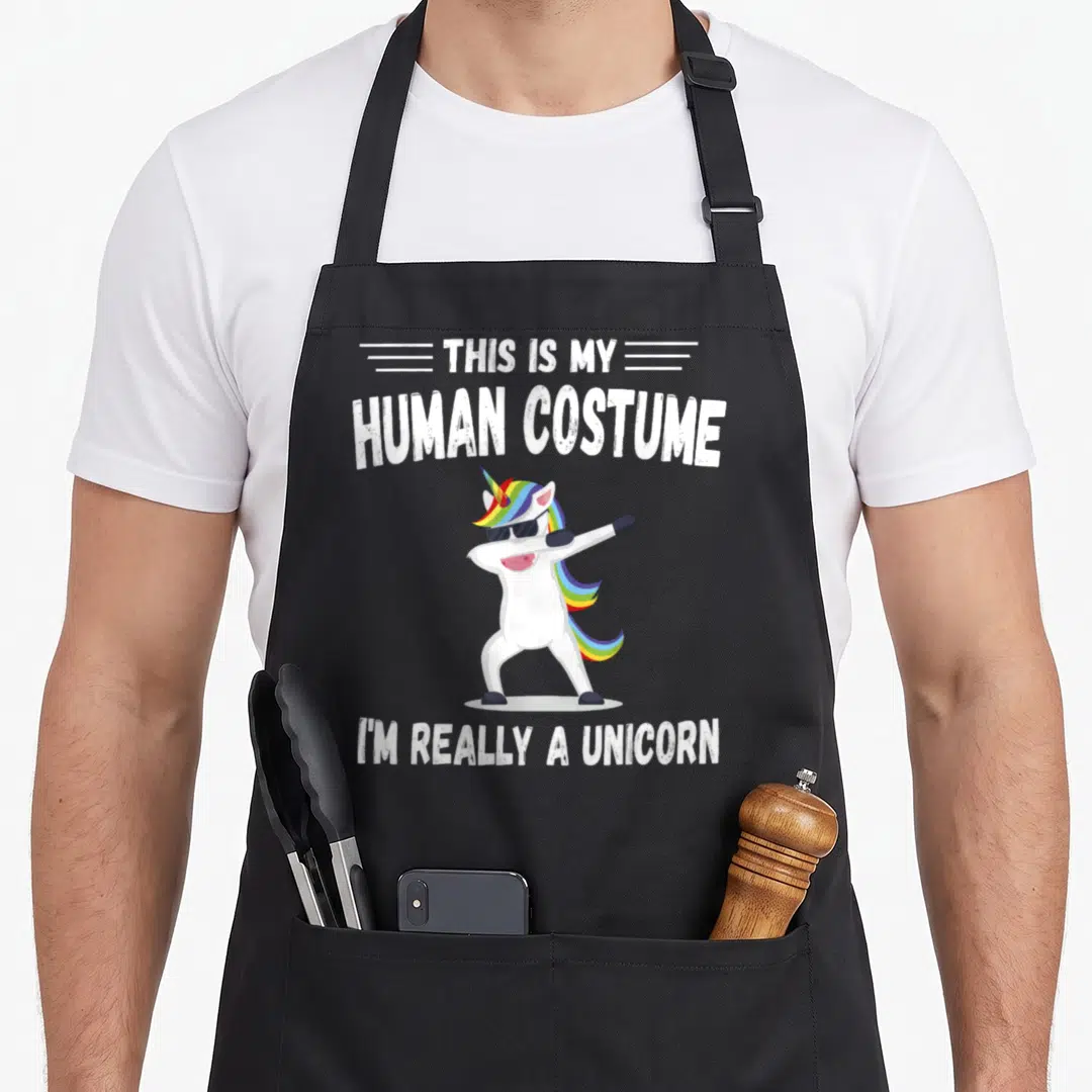 Human unicorn apron - Dabbing white unicorn with rainbow mane and sunglasses below text This Is My Human Costume I'm — shown on a black cotton canvas apron against a white background