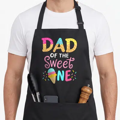 Family ice cream apron - DAD OF THE Sweet ONE text in waffle cone font with pink blue yellow sprinkles and ice cream — shown on a black cotton canvas apron against a white background