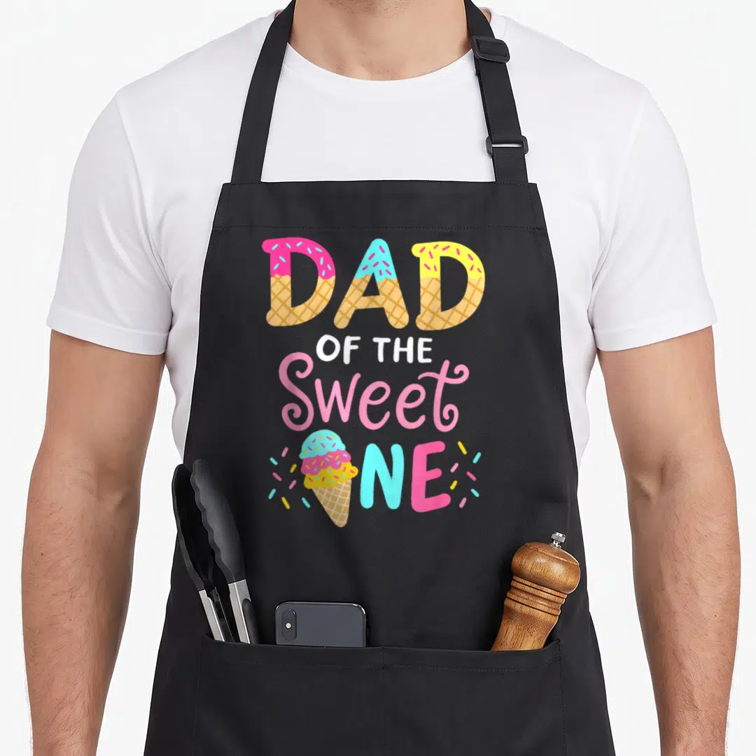 Family ice cream apron - DAD OF THE Sweet ONE text in waffle cone font with pink blue yellow sprinkles and ice cream — shown on a black cotton canvas apron against a white background