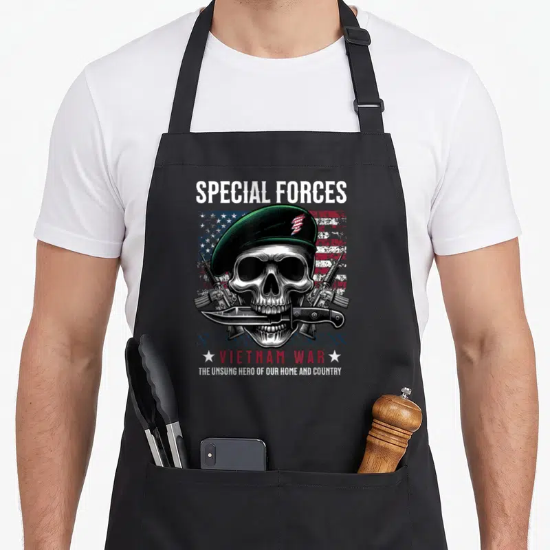Special army green apron - Skull in green beret with rifles and knife over distressed flag with Vietnam War text — shown on a black cotton canvas apron against a white background