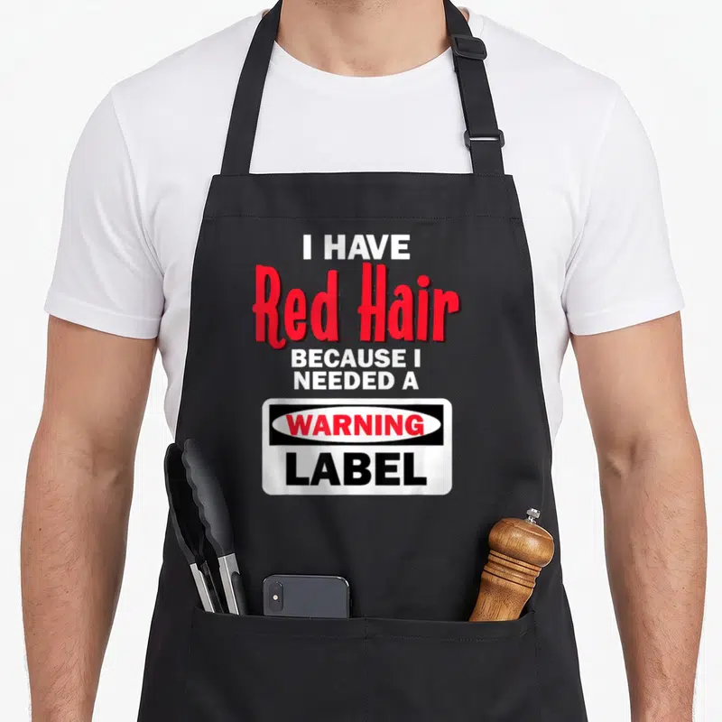 Warning hair apron - I HAVE Red Hair BECAUSE I NEEDED A WARNING LABEL text in red white black — shown on a black cotton canvas apron against a white background