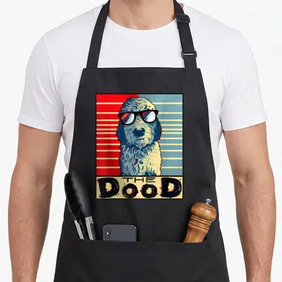 Golden goldendoodle apron - Retro dog with sunglasses and THE Dood text on red and blue stripes — shown on a black cotton canvas apron against a white background