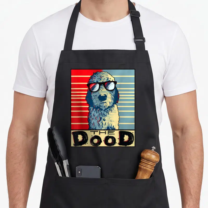 Golden goldendoodle apron - Retro dog with sunglasses and THE Dood text on red and blue stripes — shown on a black cotton canvas apron against a white background