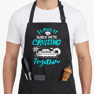 Cruising essential apron - Cyan and white I LOVE IT WHEN WE'RE CRUISING Together text with ship, palm tree, lifebuoy — shown on a black cotton canvas apron against a white background