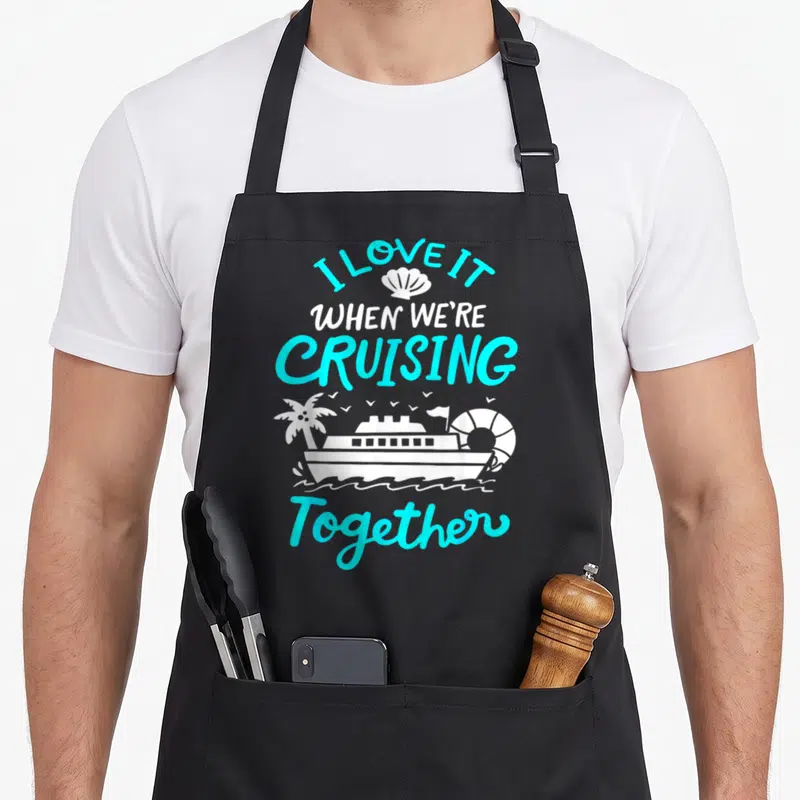 Cruising essential apron - Cyan and white I LOVE IT WHEN WE'RE CRUISING Together text with ship, palm tree, lifebuoy — shown on a black cotton canvas apron against a white background