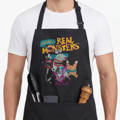 Aaahh character aprons - Four colorful monsters below yellow Real Monsters text and a blue speech bubble — shown on a black cotton canvas apron against a white background