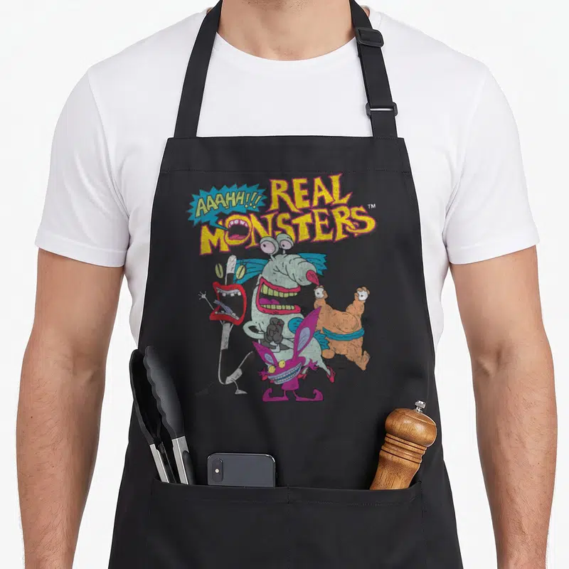 Aaahh character aprons - Four colorful monsters below yellow Real Monsters text and a blue speech bubble — shown on a black cotton canvas apron against a white background