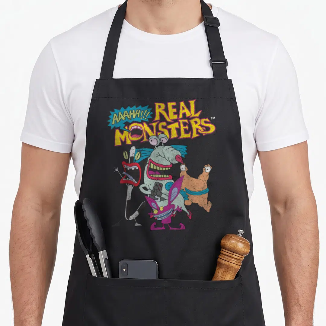 Aaahh character aprons - Four colorful monsters below yellow Real Monsters text and a blue speech bubble — shown on a black cotton canvas apron against a white background