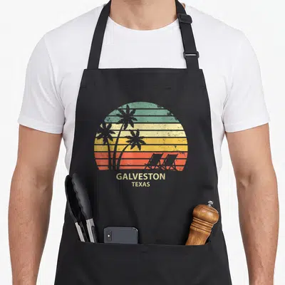 Galveston texas apron - Retro sunset with striped green yellow orange red white background, palm tree and beach chair — shown on a black cotton canvas apron against a white background