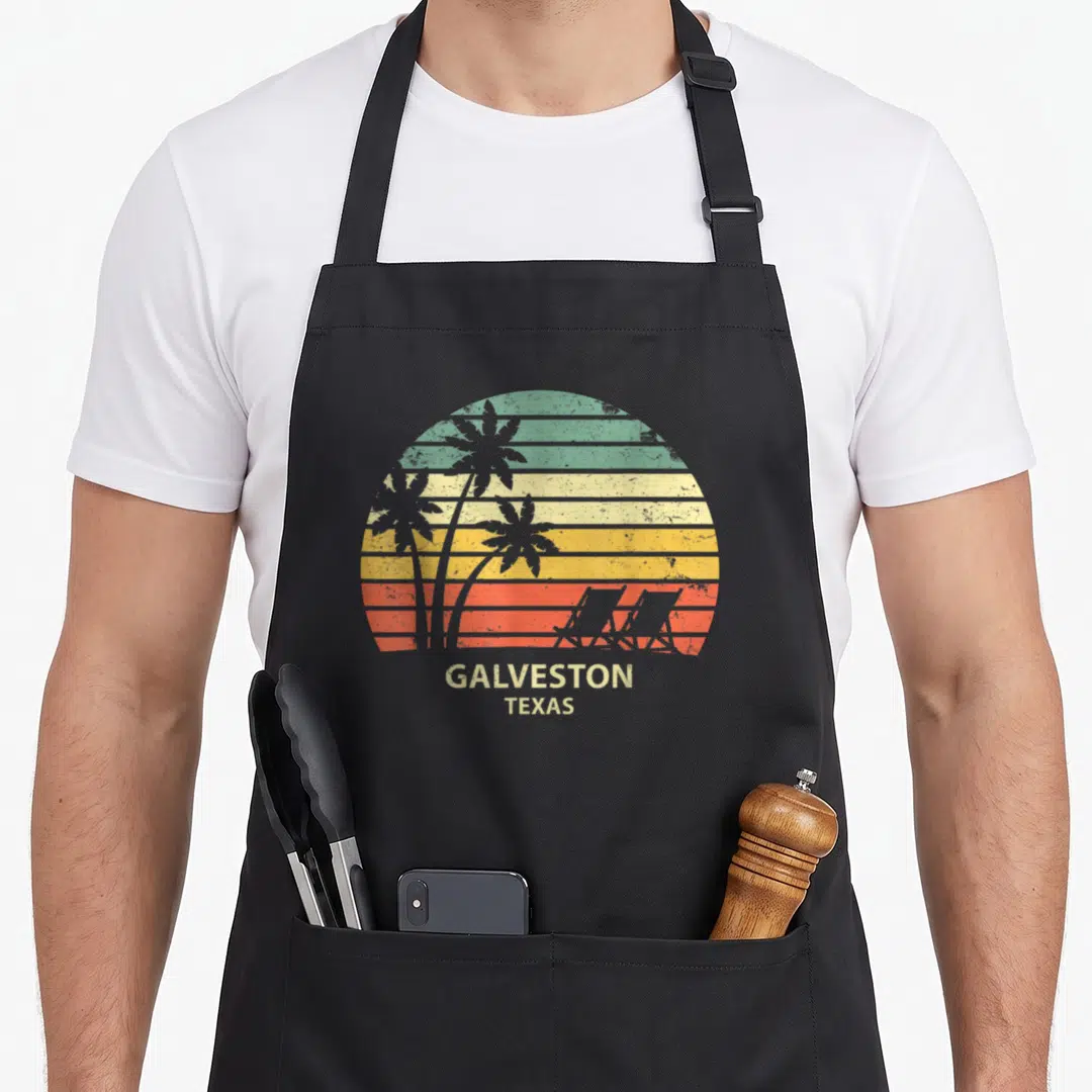 Galveston texas apron - Retro sunset with striped green yellow orange red white background, palm tree and beach chair — shown on a black cotton canvas apron against a white background