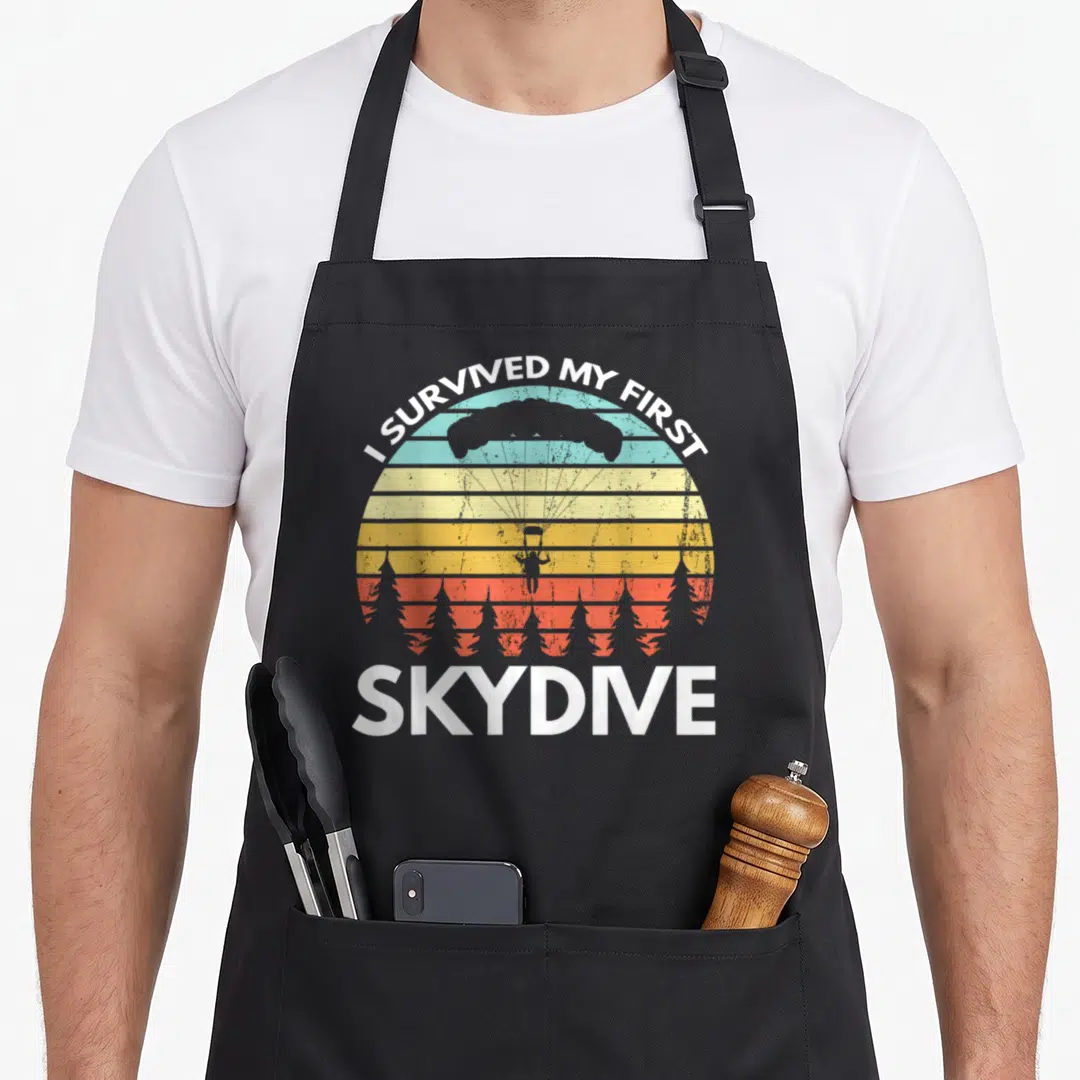 Survived skydiving apron - Retro striped sun with skydiver silhouette and white text I SURVIVED MY FIRST SKYDIVE — shown on a black cotton canvas apron against a white background