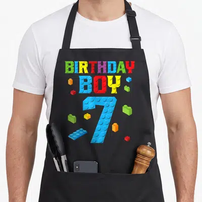 Builder building aprons - Colorful brick style BIRTHDAY BOY 7 text with scattered toy blocks in blue red yellow green — shown on a black cotton canvas apron against a white background