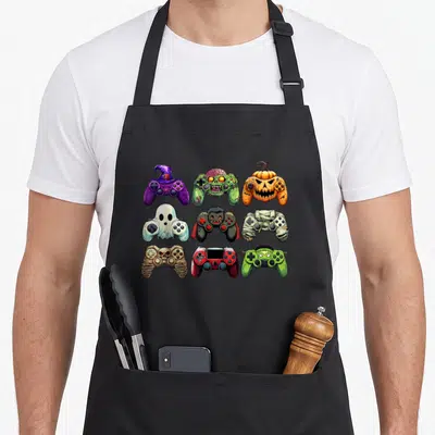Zombie mummy and me aprons - Nine monster game controllers in a 3x3 grid featuring purple witch, green zombie, orange — shown on a black cotton canvas apron against a white background