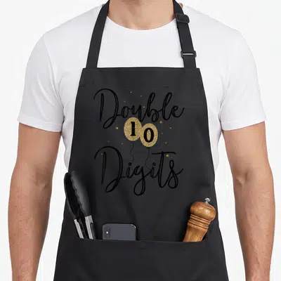 Vintage digit birthday apron - Gold balloons forming 10 with Double Digits black script and scattered stars — shown on a black cotton canvas apron against a white background