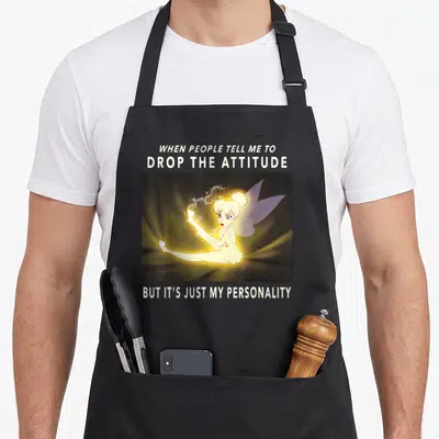 Peter meme apron - Fairy sitting with magic dust under text about dropping attitude and personality — shown on a black cotton canvas apron against a white background