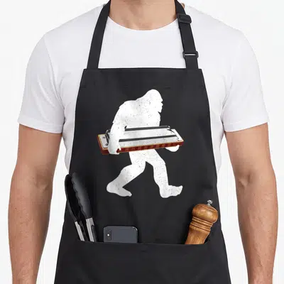 Harmonicist harmonica apron - White bigfoot silhouette walking right carrying a brown and silver harmonica — shown on a black cotton canvas apron against a white background