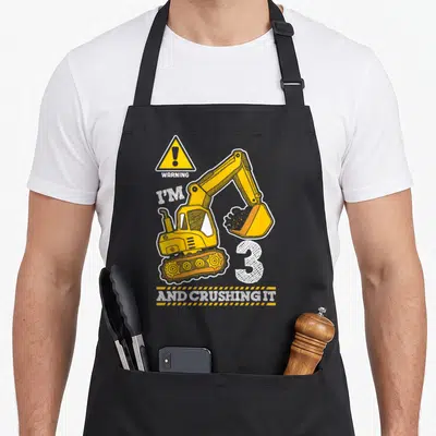 Construction truck apron - Yellow excavator graphic with WARNING I'M 3 AND CRUSHING IT text on stripes — shown on a black cotton canvas apron against a white background