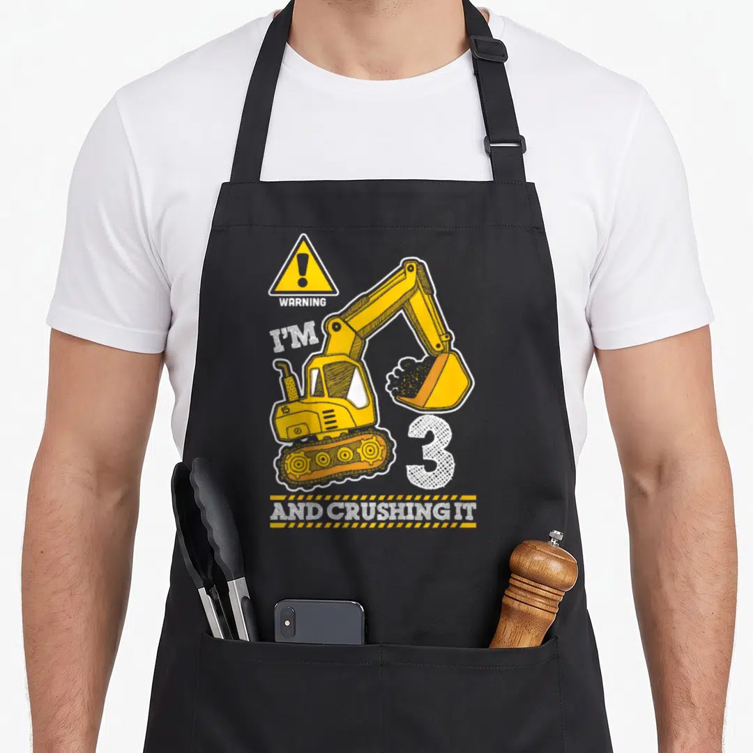 Construction truck apron - Yellow excavator graphic with WARNING I'M 3 AND CRUSHING IT text on stripes — shown on a black cotton canvas apron against a white background