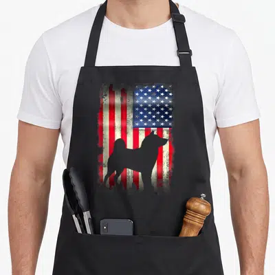 Akita dog american flag apron - Grey dog silhouette standing in front of a distressed red white and blue flag background — shown on a black cotton canvas apron against a white background
