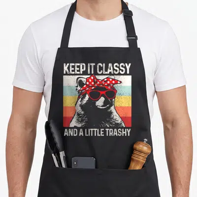 Trashy raccoon apron - Grey raccoon wearing red glasses and bandana between text KEEP IT CLASSY AND A LITTLE TRASHY — shown on a black cotton canvas apron against a white background
