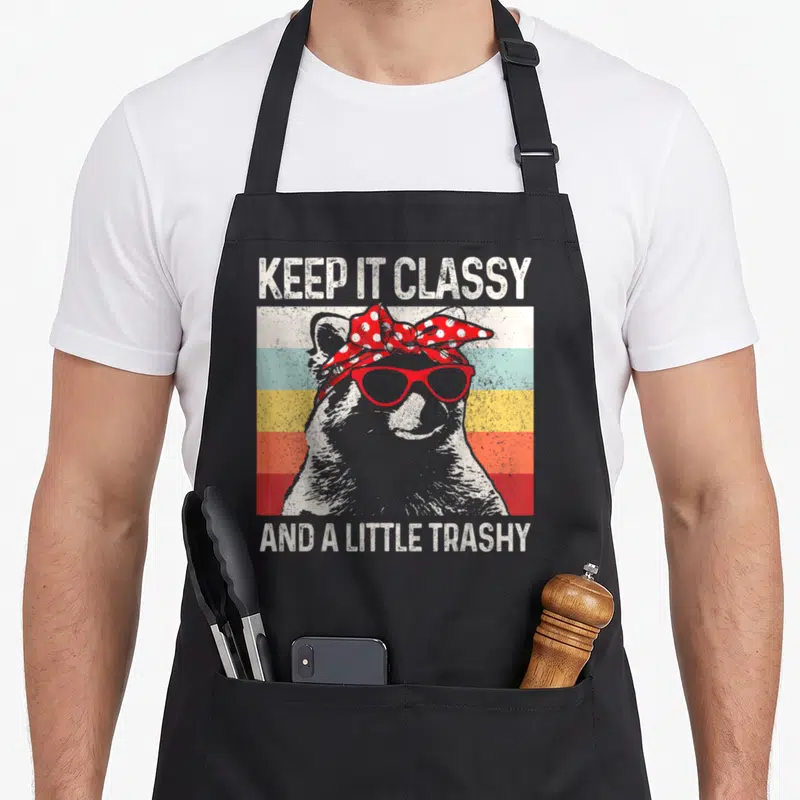 Trashy raccoon apron - Grey raccoon wearing red glasses and bandana between text KEEP IT CLASSY AND A LITTLE TRASHY — shown on a black cotton canvas apron against a white background