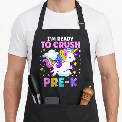 Preschool unicorn apron - Cartoon unicorn on cloud with I'M READY TO CRUSH PRE-K text in pink blue yellow purple white — shown on a black cotton canvas apron against a white background
