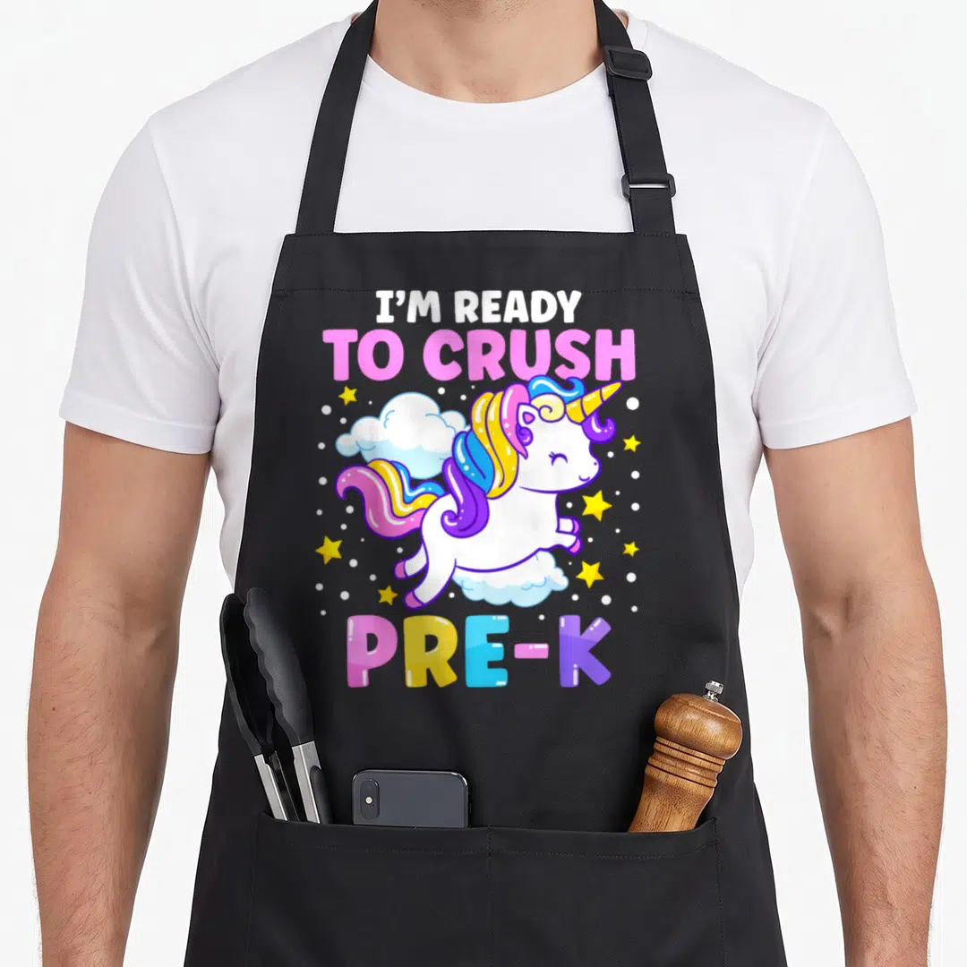 Preschool unicorn apron - Cartoon unicorn on cloud with I'M READY TO CRUSH PRE-K text in pink blue yellow purple white — shown on a black cotton canvas apron against a white background