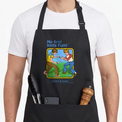 Steven knife apron - Cartoon two boys fighting with knives under My first Knife Fight text — shown on a black cotton canvas apron against a white background