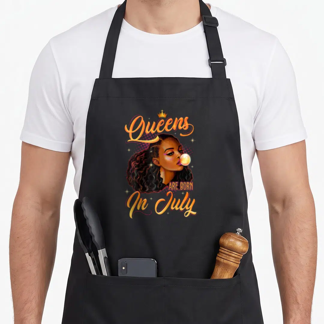 July cancer birthday apron - Queens ARE BORN In July text with crown and cartoon woman blowing bubble in orange — shown on a black cotton canvas apron against a white background