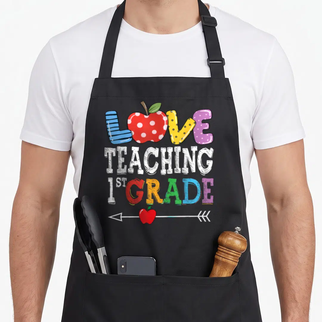 Teaching art teacher apron - Colorful LOVE TEACHING 1ST GRADE text with striped and dotted letters and red apples — shown on a black cotton canvas apron against a white background