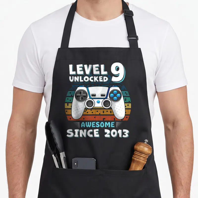 9yr birthday apron - Game controller with level 9 unlocked text on pixelated background — shown on a black cotton canvas apron against a white background