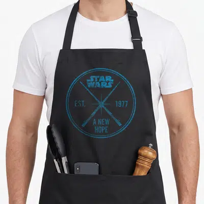 Hope craft apron - Blue circular stamp design with STAR WARS EST 1977 A NEW HOPE text and crossing lines — shown on a black cotton canvas apron against a white background