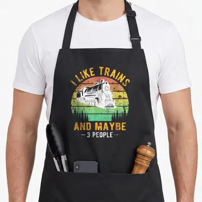 Railroad model apron - White steam train in retro striped sun circle with I Like Trains text — shown on a black cotton canvas apron against a white background
