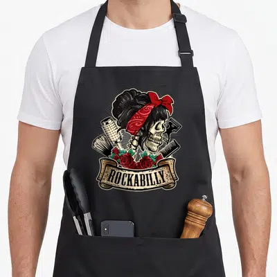 Hairstylist rockabilly apron - Skeleton with red bandana, hairbrush, comb, and roses under beige ROCKABILLY banner — shown on a black cotton canvas apron against a white background