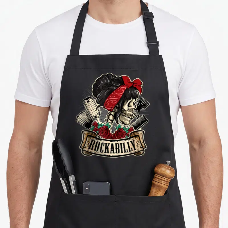 Hairstylist rockabilly apron - Skeleton with red bandana, hairbrush, comb, and roses under beige ROCKABILLY banner — shown on a black cotton canvas apron against a white background