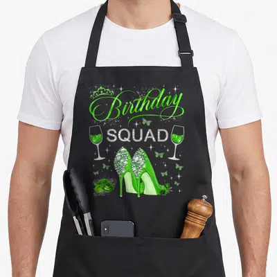 Diva butterfly apron - Green high heels andBirthday SQUAD text with butterflies and champagne glasses — shown on a black cotton canvas apron against a white background