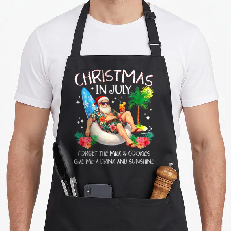 Beach christmas tree apron - Santa Claus sitting in bean bag with surfboard and drink under Christmas In July text — shown on a black cotton canvas apron against a white background