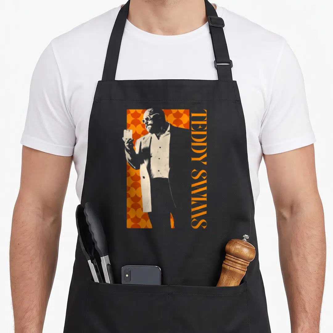 Teddy milk aprons - Beige man silhouette holding glass against orange scallops with vertical TEDDY SWIMS text — shown on a black cotton canvas apron against a white background