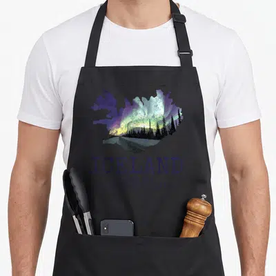 Map icelandic apron - Retro map of Iceland silhouette filled with green aurora and pine trees above dark purple text — shown on a black cotton canvas apron against a white background