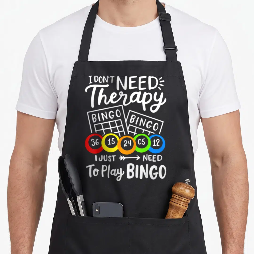 Therapie bingo apron - I DON'T NEED Therapy BINGO text with two cards and five red yellow orange green blue numbered — shown on a black cotton canvas apron against a white background
