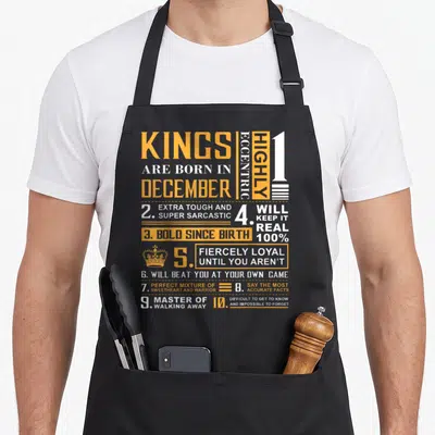 December king apron - Yellow and white text listing ten sarcastic traits for December kings with a small crown icon — shown on a black cotton canvas apron against a white background