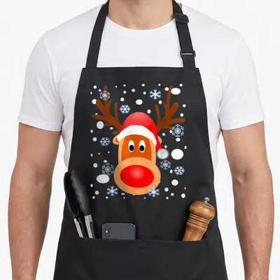 Snow reindeer christmas apron - Brown cartoon reindeer head with red Santa hat and white snowflakes — shown on a black cotton canvas apron against a white background