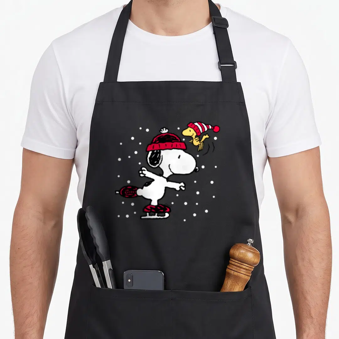 Woodstock skate holiday apron - Snoopy skating in red hat and boots with Woodstock and white snowflakes — shown on a black cotton canvas apron against a white background