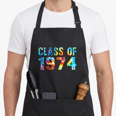 Class reunion tie-dye apron - CLASS OF 1974 text filled with blue yellow red green white tie-dye pattern — shown on a black cotton canvas apron against a white background