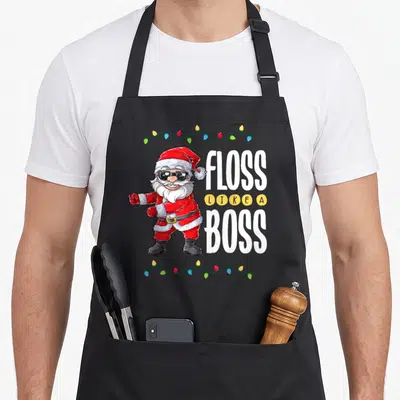 Santa boss apron - Cartoon Santa flossing with sunglasses next to stacked text FLOSS LIKE A BOSS framed by Christmas — shown on a black cotton canvas apron against a white background