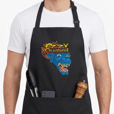 Ozzy tattoo apron - Blue demon head with OZZY OSBOURNE text and yellow orange red flames — shown on a black cotton canvas apron against a white background