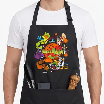 Monstar jam apron - Orange basketball with SPACE JAM text and cartoon characters in purple green yellow orange — shown on a black cotton canvas apron against a white background