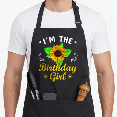 Theme sunflower apron - I'M THE Birthday Girl text with yellow sunflower and white swirls — shown on a black cotton canvas apron against a white background