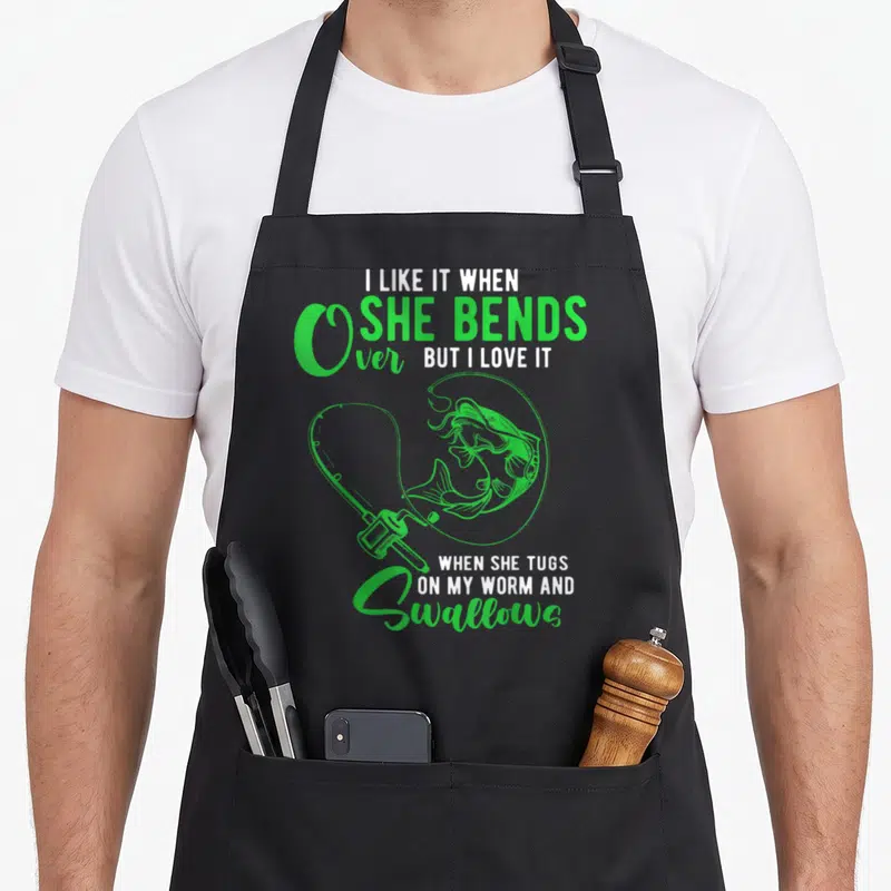 Cute she fishing aprons - Green and white text reading I like it when she bends over with line art fish and rod — shown on a black cotton canvas apron against a white background